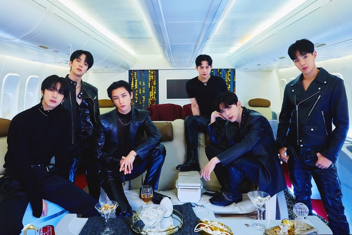 비행기 내부에서 가죽과 검은색 옷을 입은 여섯 명의 남성이 소파와 의자에 앉아 포즈를 취하고 있다. 테이블 위에는 술잔과 장식품이 놓여 있다.