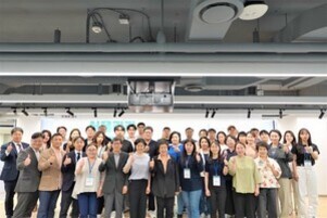 실내에서 여러 명의 남녀가 단체로 서서 기념사진을 찍고 있다. 대부분 엄지손가락을 들어 올리고 있으며, 밝은 분위기이다.