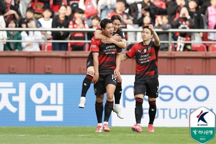 축구 경기장에서 포항 스틸러스 선수들이 골을 넣고 기뻐하며 서로를 축하하고 있다. 관중석에는 많은 팬들이 응원하고 있다. 오른쪽 아래에는 K리그 로고가 보인다.