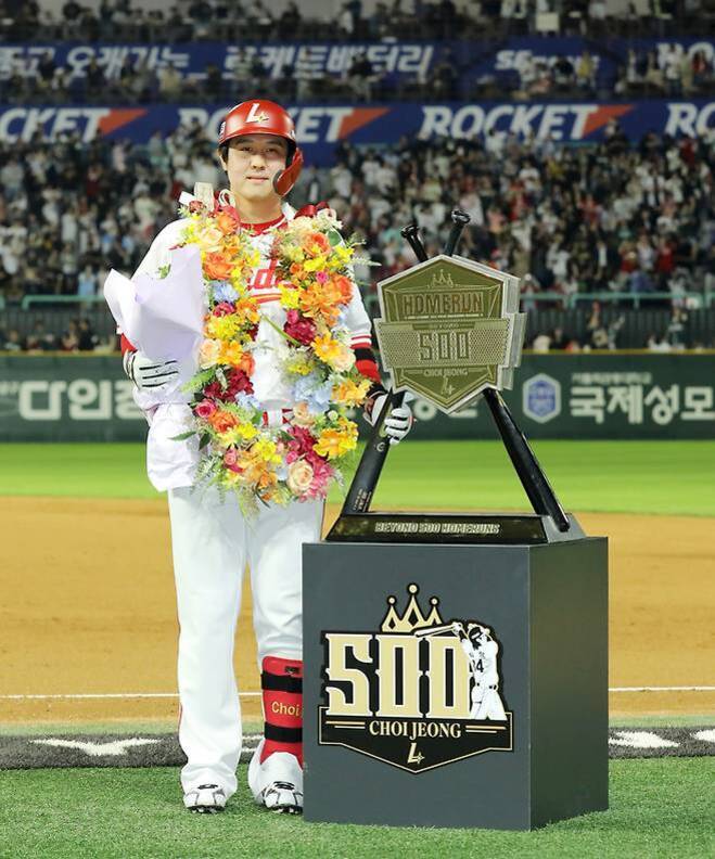 야구 경기장에서 꽃목걸이와 헬멧을 쓴 야구 선수가 500홈런 기념 트로피 옆에 서서 꽃다발을 들고 있다. 뒤에는 관중석이 보인다.