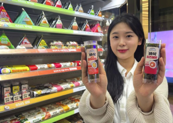 편의점 진열대 앞에서 삼각김밥과 다양한 간편식을 배경으로 한 여성이 바삭치즈볼과 바삭참치마요 제품을 두 손에 들고 미소 짓고 있다.