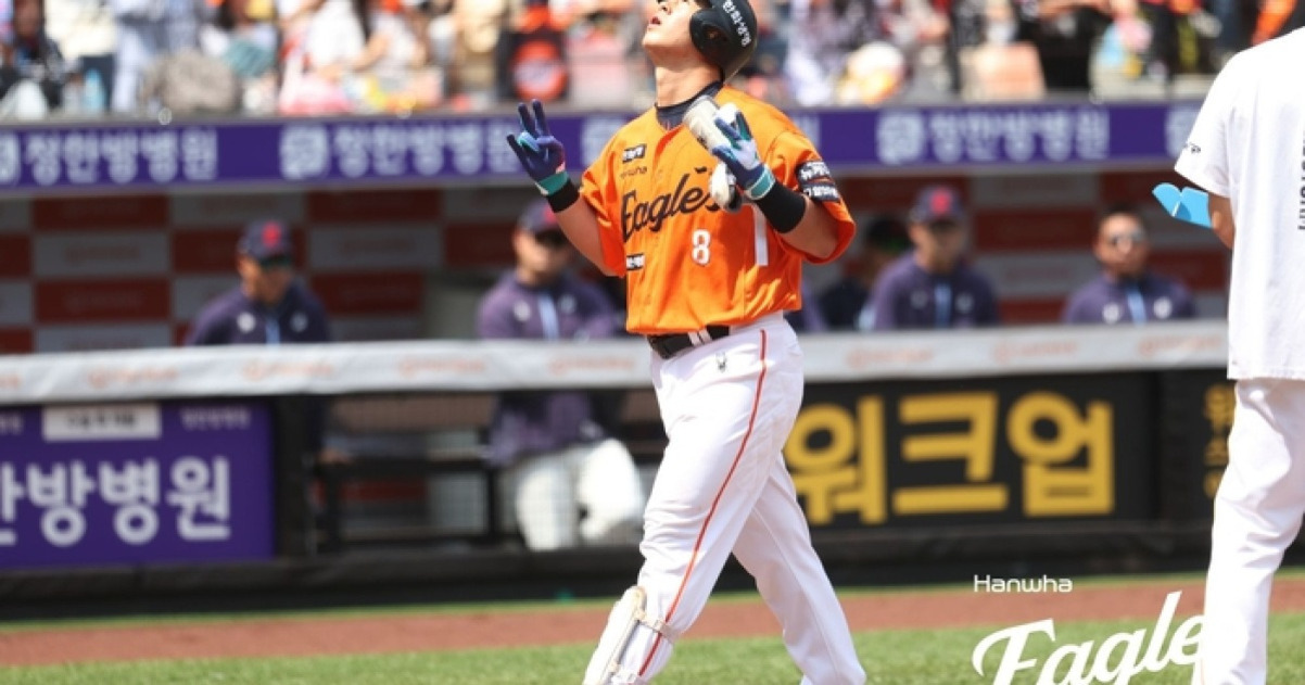 야구 경기장에서 한화 이글스 유니폼을 입은 야구 선수가 헬멧을 쓰고 두 손가락으로 브이 표시를 하며 하늘을 올려다보고 있다. 뒤에는 덕아웃과 관중들이 보인다.