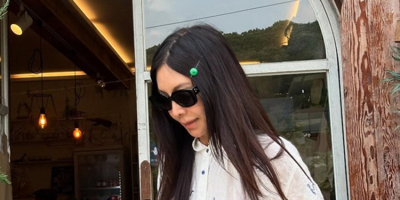 긴 생머리에 선글라스를 쓴 여성이 흰 셔츠를 입고 문 앞에 서 있다.