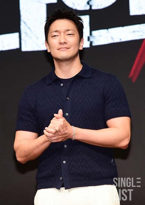 짧은 소매의 네이비색 셔츠와 흰 바지를 입은 남성이 두 손을 모으고 무대 위에 서 있다.