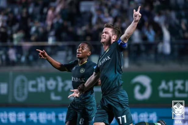 축구 경기장에서 두 명의 축구 선수가 골을 넣은 후 기뻐하며 세리머니를 하고 있다. 한 선수는 팔을 벌리고, 다른 선수는 손가락으로 하늘을 가리키고 있다. 관중들이 뒤에 보인다.