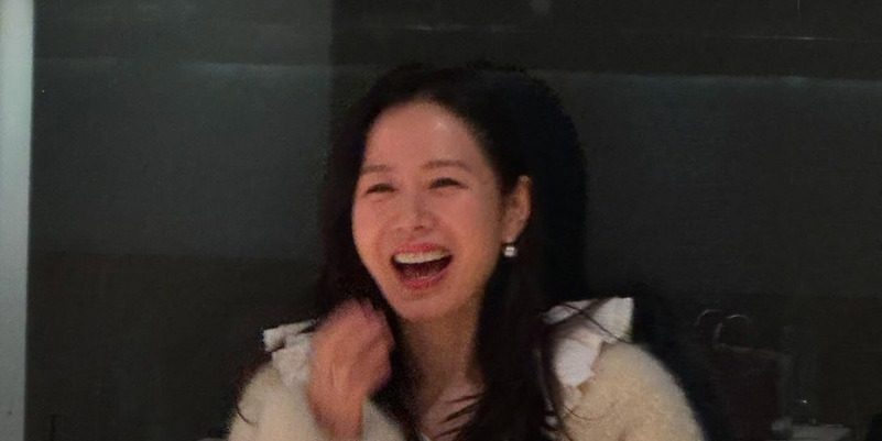 흰색 옷을 입은 여성이 밝게 웃으며 손으로 입을 가리고 있다.