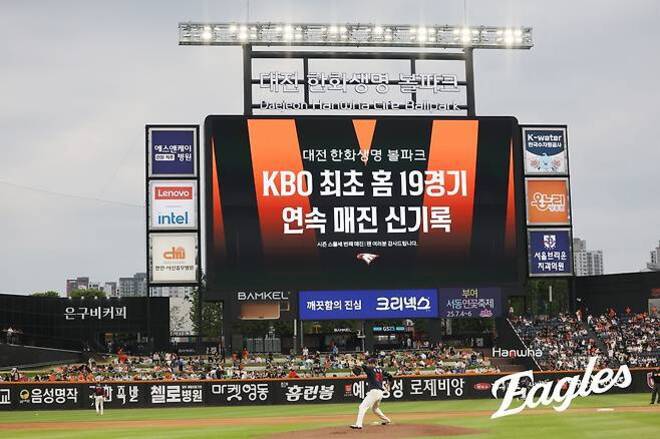 대전 한화생명 이글스파크 야구장에서 전광판에 'KBO 최초 홈 19경기 연속 매진 신기록'이라는 문구가 표시되어 있고, 경기장에는 관중들이 앉아 있으며 선수들이 경기를 준비하고 있다.
