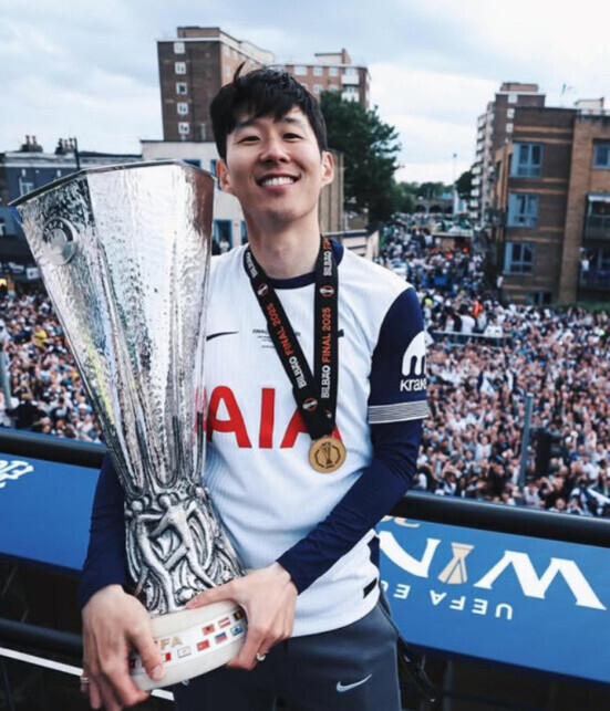 축구 유니폼을 입은 남성이 큰 트로피를 들고 메달을 목에 건 채 미소 짓고 있으며, 뒤에는 많은 인파가 모여 있는 야외 거리 풍경이 보인다.
