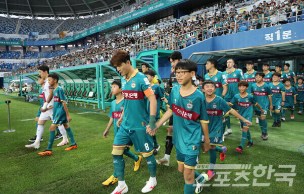 축구 경기장에서 선수들과 어린이들이 손을 잡고 입장하는 모습, 관중석에는 많은 관중이 앉아 있다.