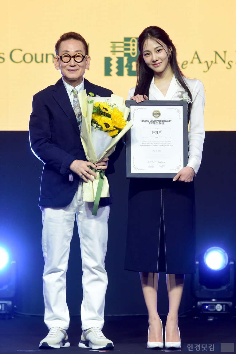 무대 위에서 한 남성과 여성이 함께 서 있다. 남성은 선글라스를 끼고 꽃다발을 들고 있으며, 여성은 상장을 들고 미소 짓고 있다. 둘 다 단정한 복장을 하고 있으며, 배경에는 노란색 스크린이 보인다.