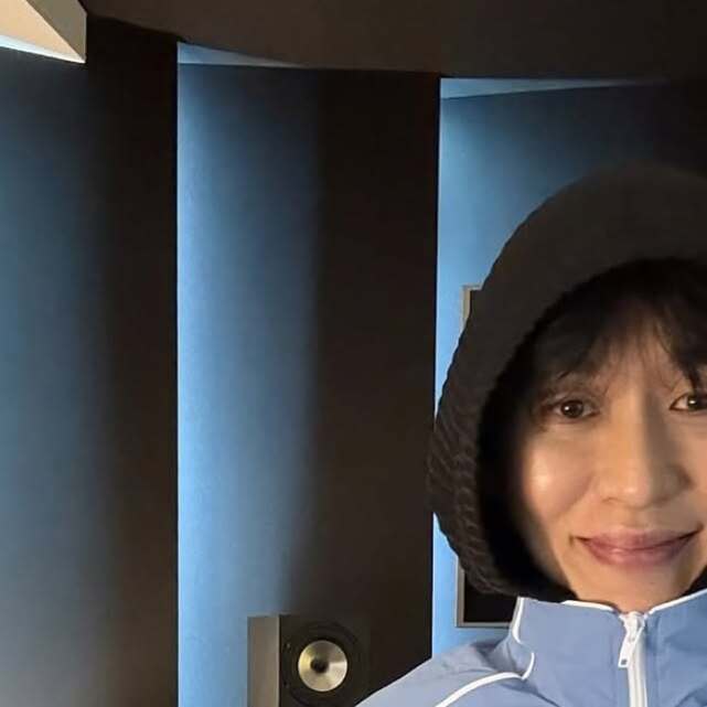 후드를 쓴 사람이 밝게 미소 짓고 있는 실내 사진의 오른쪽 반만 보임. 배경에는 스피커가 있음.