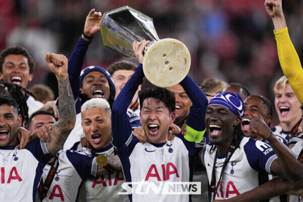 축구 선수들이 'AIA'가 적힌 유니폼을 입고 트로피를 들어 올리며 기뻐하는 모습. 중앙의 선수가 트로피를 머리 위로 들고 있고, 팀원들이 함께 환호하고 있다.