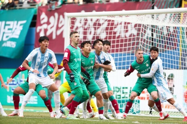 축구 경기에서 여러 명의 선수들이 골문 앞에서 공을 주시하며 수비와 공격을 준비하고 있다.