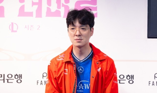 안경을 쓴 남성이 파란색 유니폼과 빨간색 점퍼를 입고 실내에서 정면을 바라보고 있다.