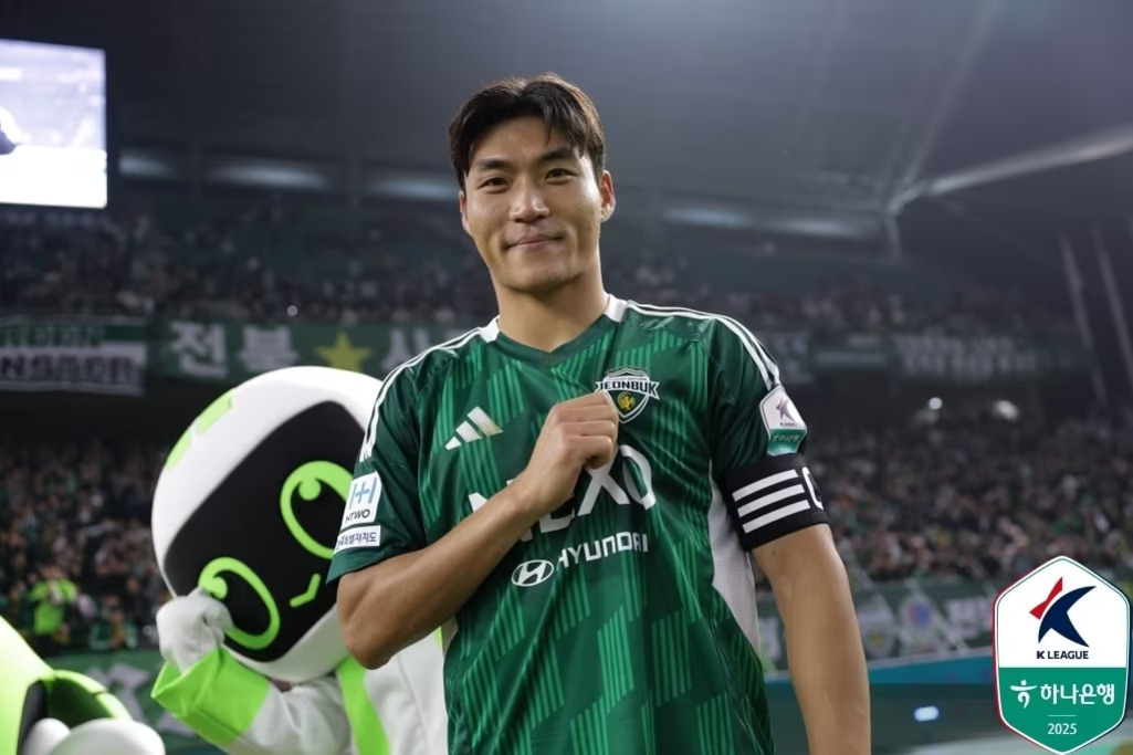 축구 경기장에서 전북현대 유니폼을 입은 선수가 가슴에 손을 얹고 미소를 짓고 있으며, 뒤에는 마스코트와 관중이 보인다.