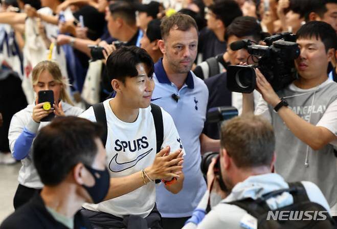 한 남성이 'SPURS'라고 적힌 흰색 티셔츠를 입고 공항에서 미소를 지으며 손뼉을 치는 모습. 주변에는 많은 사람들이 카메라와 휴대폰으로 촬영하고 있다.