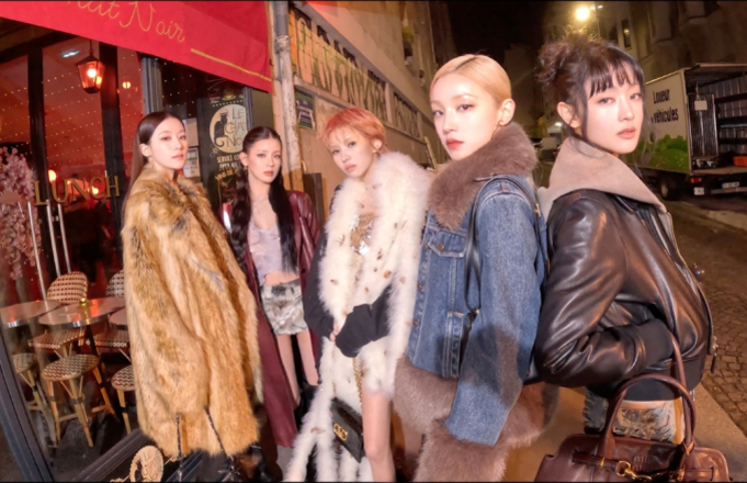 다섯 명의 여성이 밤거리에서 화려한 겨울 옷차림으로 포즈를 취하고 있다. 배경에는 카페와 테이블이 보인다.