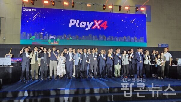 여러 명의 사람들이 무대 위에 서서 단체 사진을 찍고 있으며, 배경에는 'PlayX4'라는 큰 파란색 간판이 걸려 있다.
