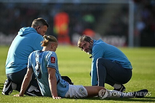 축구 경기장에서 한 선수가 잔디 위에 앉아 있고, 두 명의 트레이너가 곁에서 상태를 살피고 있다.