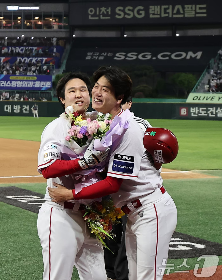 야구 경기장에서 SSG 랜더스 유니폼을 입은 두 명의 선수가 꽃다발을 들고 서로를 포옹하며 기뻐하는 모습