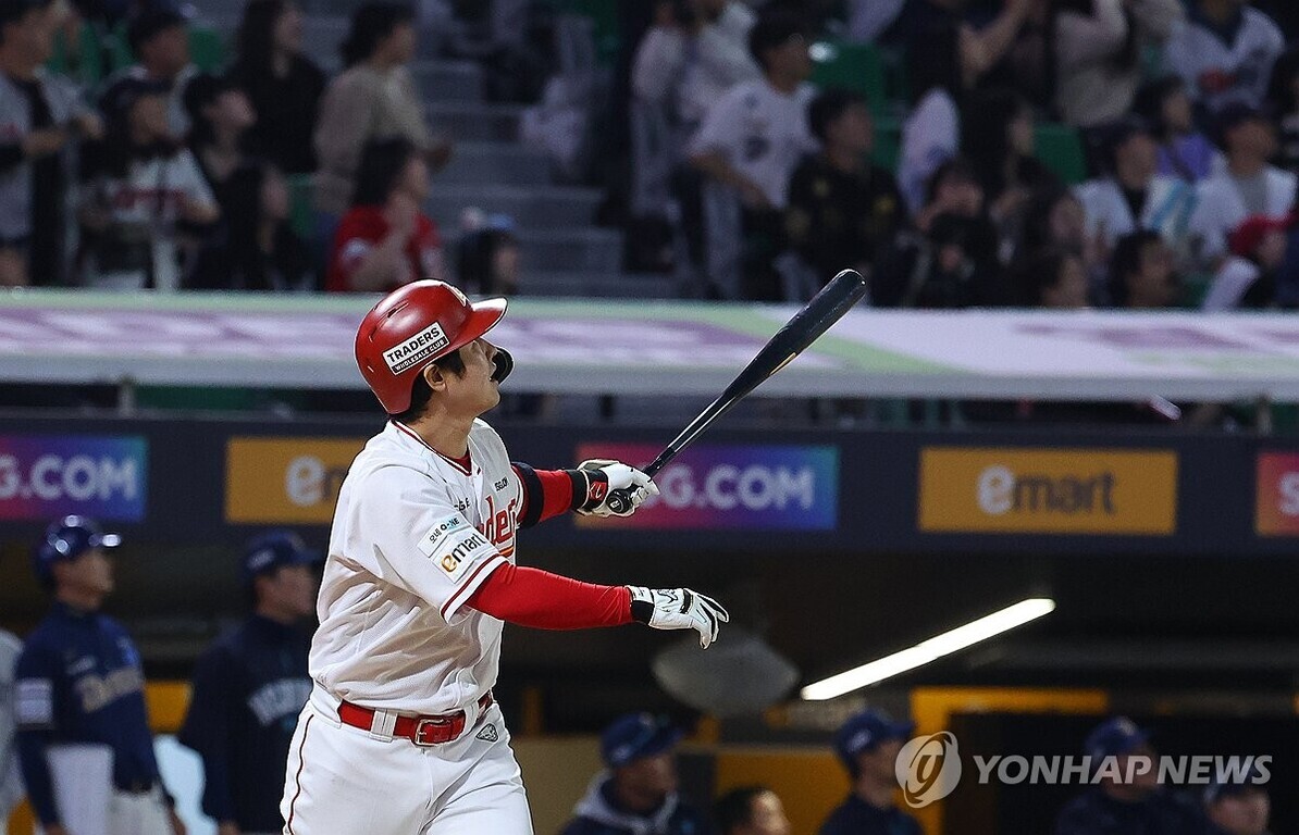야구 선수가 헬멧과 유니폼을 착용하고 방망이를 휘두른 후 타구를 바라보고 있다. 관중석에는 사람들이 앉아 경기를 지켜보고 있다.