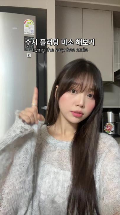 긴 머리를 가진 젊은 여성이 부엌에서 카메라를 향해 손가락을 들어 올리고 있다. 상단에는 '수지 플러팅 미소 해보기'라는 자막이 있다.