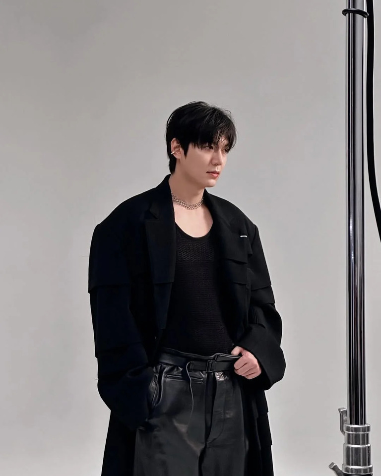 검정색 긴 코트와 가죽 바지를 입은 남성이 실내 스튜디오에서 포즈를 취하고 있다. 배경은 심플한 회색이며, 조명 장비 일부가 오른쪽에 보인다.