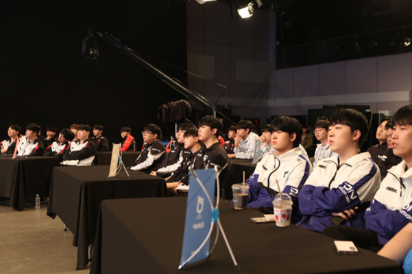 여러 명의 젊은 남성들이 팀 유니폼을 입고 실내 행사장 테이블에 앉아 무언가를 집중해서 바라보고 있다.