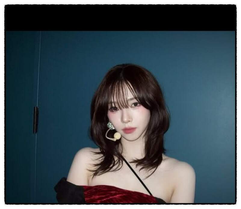 어깨를 드러낸 의상을 입고 무대용 헤드셋 마이크를 착용한 긴 머리 여성의 상반신 사진. 배경은 파란색 벽이다.