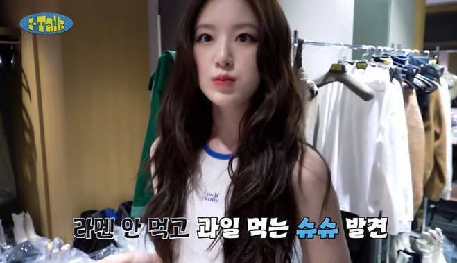 긴 머리의 젊은 여성이 흰색 민소매 상의를 입고 실내에서 카메라를 바라보고 있다. 화면 하단에는 '라멘 안 먹고 과일 먹는 슈슈 발견'이라는 자막이 있다. 뒤에는 옷걸이에 여러 옷들이 걸려있다.