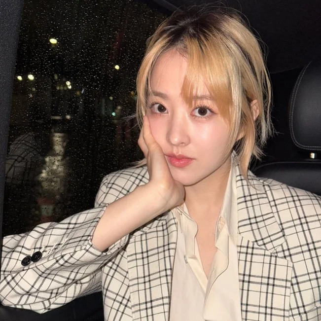 체크무늬 재킷을 입은 여성이 차 안에서 손으로 턱을 괴고 카메라를 바라보고 있다. 창밖에는 비가 내리고 있다.