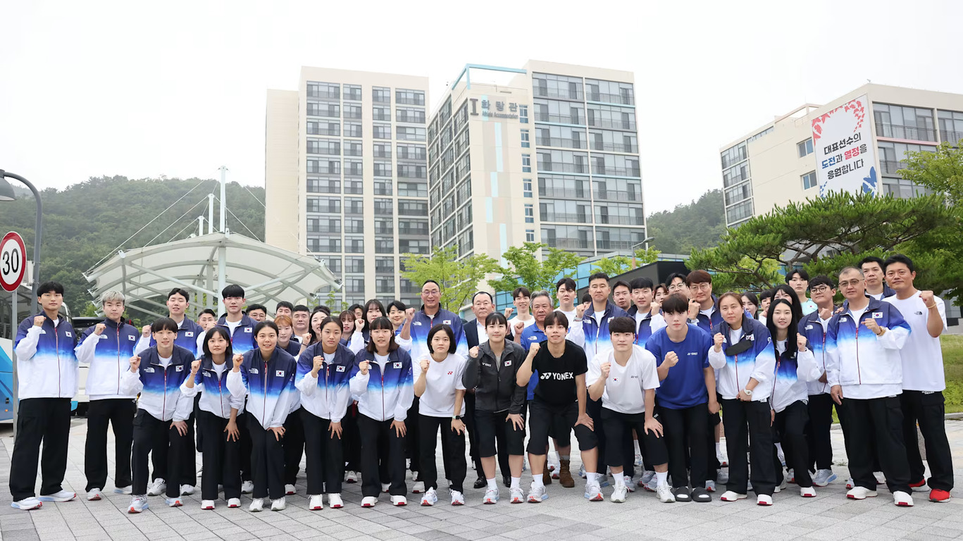 대한민국 국가대표 선수단이 단체로 유니폼을 입고 건물 앞에서 파이팅 포즈를 취하며 기념 촬영을 하고 있다.