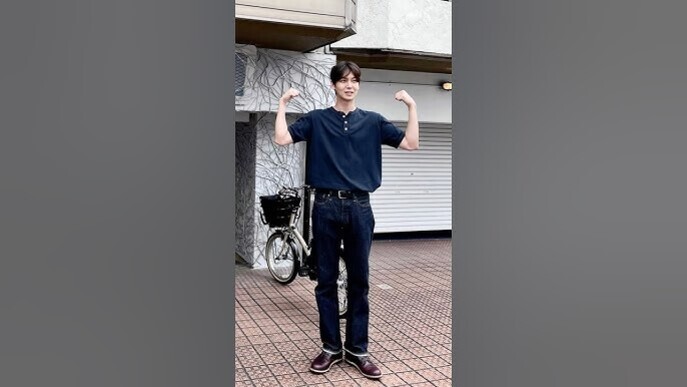 남성이 야외에서 팔을 들어 근육을 자랑하는 포즈를 취하고 있다. 뒤에는 자전거와 닫힌 셔터가 보인다.