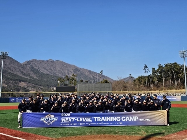 야구 경기장에서 단체로 모여 있는 선수들과 코치들이 '2025 NEXT-LEVEL TRAINING CAMP IN GJANG'이라고 적힌 현수막 뒤에서 포즈를 취하고 있다. 배경에는 산과 나무가 보인다.