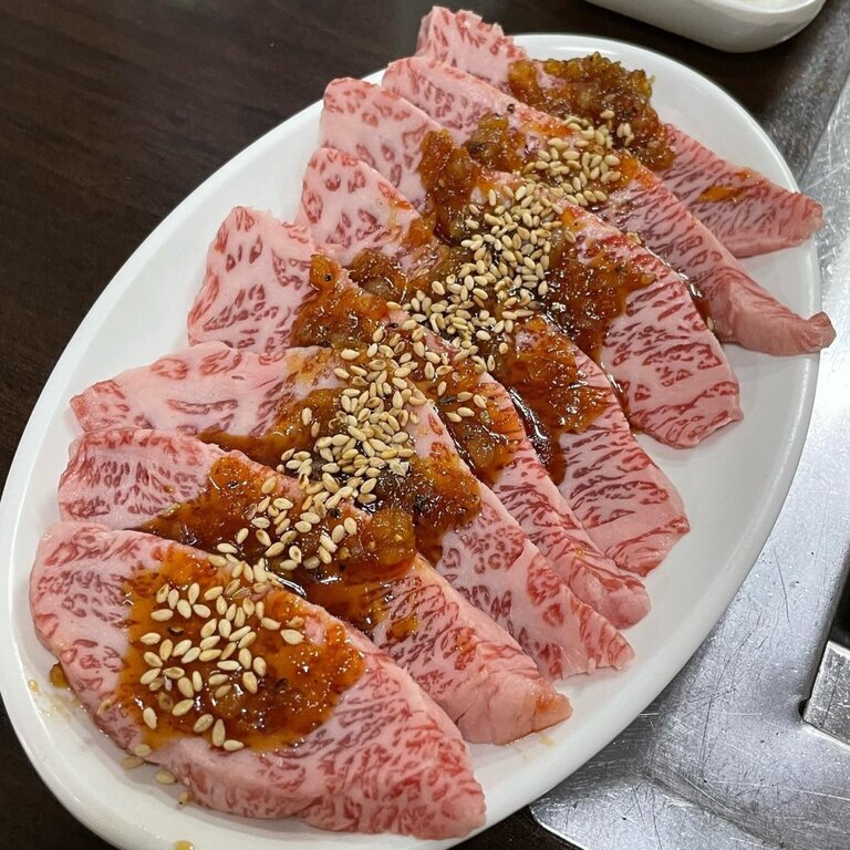 흰 접시에 마블링이 풍부한 생 소고기 구이용 고기가 가지런히 놓여 있고, 위에는 양념장과 참깨가 뿌려져 있다.