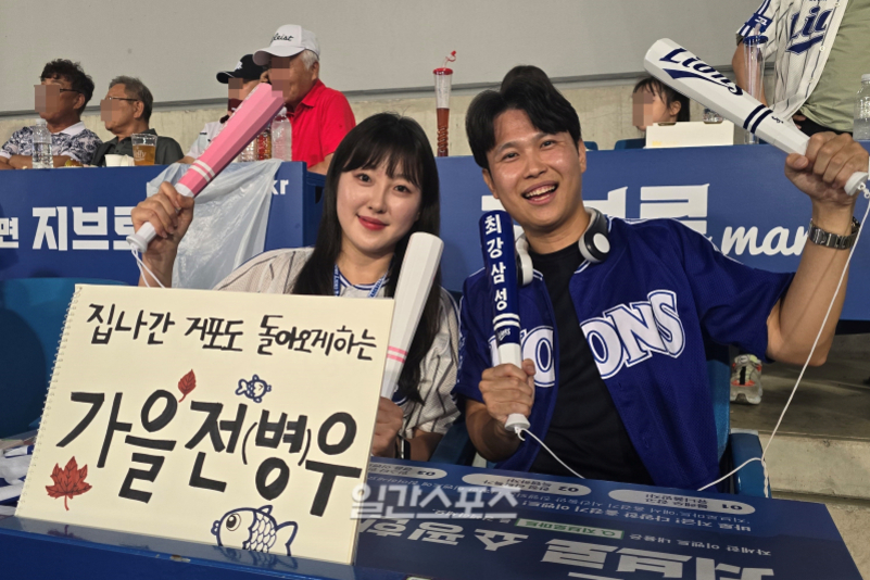 야구 경기장에서 삼성 라이온즈 유니폼을 입은 남녀가 응원도구를 들고 미소 짓고 있다. 여성은 '집나간 잭또 돌아오게 하는 가을전병우'라는 문구가 적힌 피켓을 들고 있다.