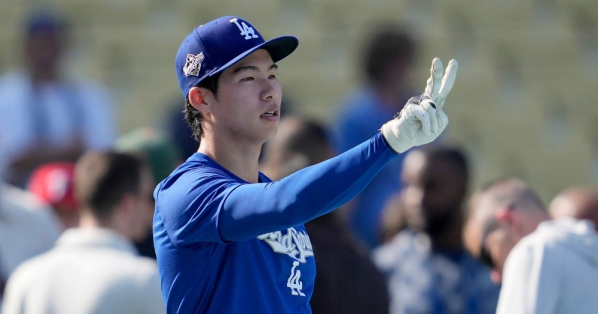 파란색 LA 다저스 유니폼과 모자를 쓴 야구 선수가 흰 장갑을 끼고 손가락으로 브이(V)자를 만들고 있다. 배경에는 여러 사람들이 흐릿하게 보인다.