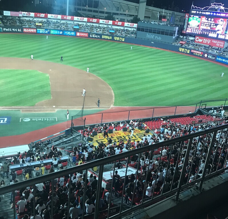 야구 경기장에 많은 관중이 앉아 있고, 밤에 경기가 진행 중이다. 내야와 외야에는 선수들이 포지션을 잡고 있으며, 전광판과 광고판이 보인다.