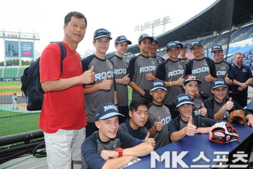 야구장 내에서 한 남성과 야구 유니폼을 입은 여러 명의 젊은 선수들이 함께 모여 엄지를 치켜세우며 단체 사진을 찍고 있다. 앞쪽에는 야구 글러브가 테이블 위에 놓여 있고, 배경에는 관중석과 경기장이 보인다.
