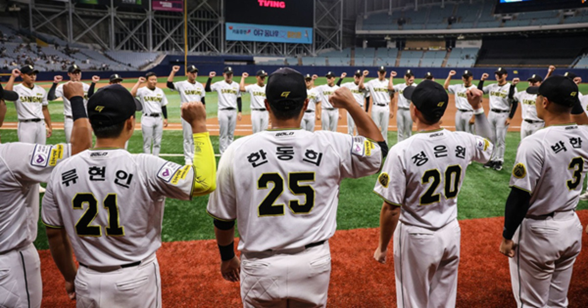 야구 유니폼을 입은 선수들이 야구장 그라운드에 원을 이루고 서서 오른팔을 들어 올리고 있다.