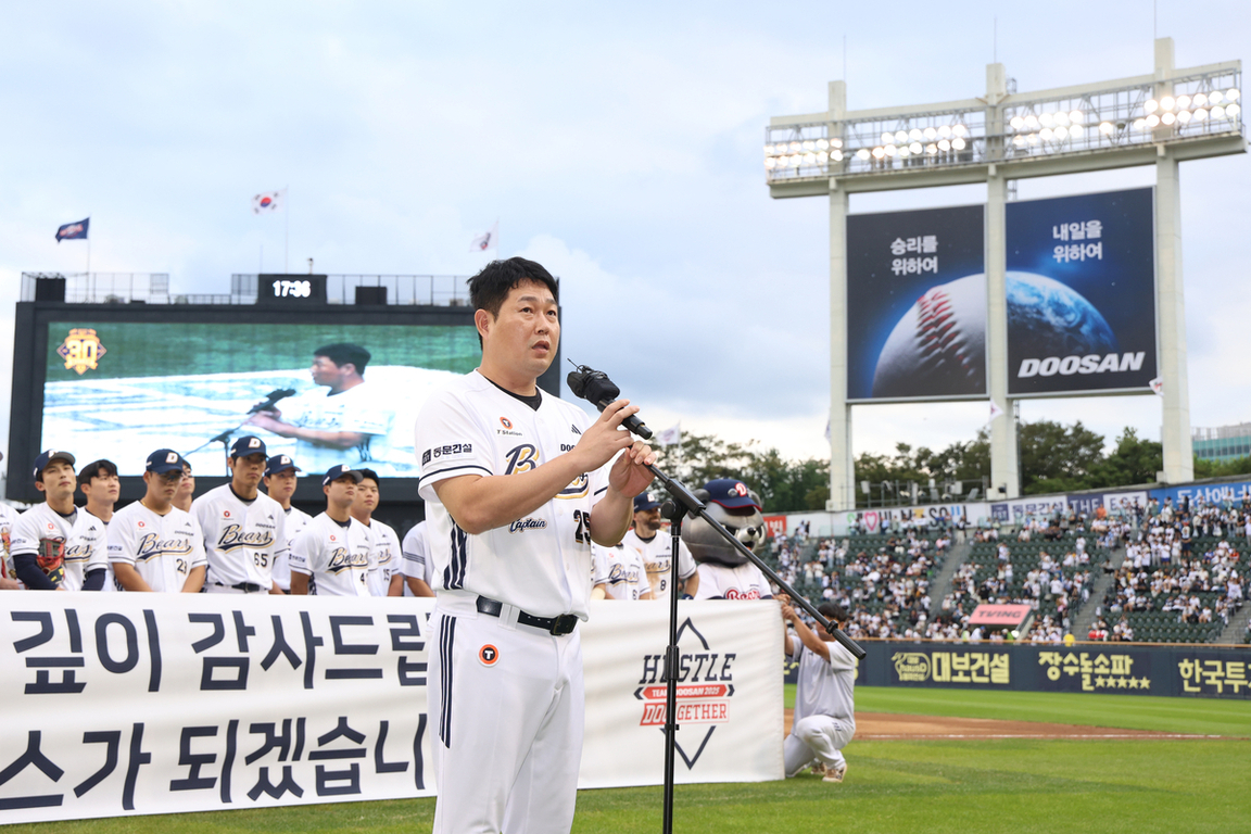 야구 경기장에서 두산 베어스 선수들이 단체로 서 있고, 한 선수가 마이크를 들고 연설하고 있다. 뒤에는 대형 전광판과 광고판, 관중석이 보인다.