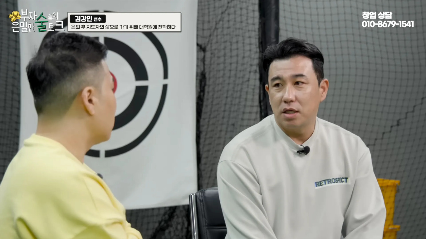 두 남성이 실내에서 대화를 나누고 있다. 한 명은 노란색 옷을 입었고, 다른 한 명은 'RETROSPECT'가 적힌 흰색 옷을 입었다. 배경에는 과녁 무늬와 그물망이 보인다. 화면 상단에는 자막과 연락처가 표시되어 있다.