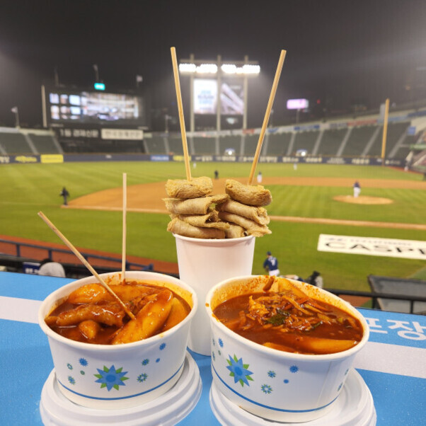 야구 경기장 관중석에서 떡볶이, 오뎅, 국물이 담긴 컵 음식 세 개가 테이블 위에 놓여 있다. 야구장이 배경으로 보인다.