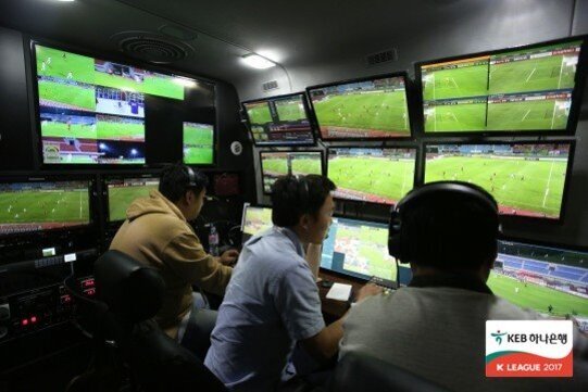 여러 명의 운영자가 헤드셋을 착용하고 축구 경기를 여러 각도에서 보여주는 모니터 화면 앞에 앉아 있는 VAR 판독실 내부 모습.