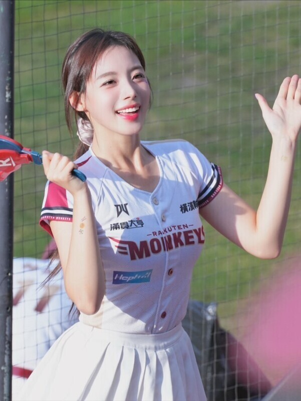 야구장 그물망 앞에서 라쿠텐 몽키스 유니폼을 입고 응원도구를 든 여성 치어리더가 미소를 지으며 손을 흔들고 있다.