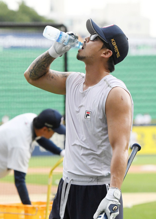 야구 연습장에 있는 남성이 모자를 쓰고 민소매 셔츠를 입은 채 물을 마시고 있다. 한 손에는 야구 배트를 들고 있으며, 뒤편에는 다른 선수가 훈련 중이다.