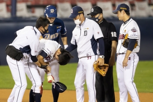 야구 경기 중 한 선수가 부상을 당해 팀 동료들과 트레이너의 도움을 받고 있다. 주변에 심판과 다른 선수들도 함께 모여 있다.