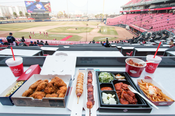 야구장 관중석에서 치킨, 꼬치, 감자튀김, 음료 등 다양한 음식이 테이블에 놓여 있고, 그 뒤로 야구 경기장이 보인다.