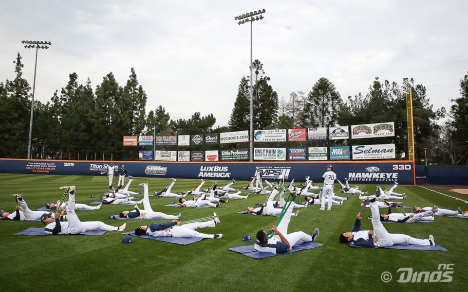 야구 선수들이 야외 경기장 잔디 위에서 스트레칭 운동을 하고 있다. 대부분 누워서 다리를 들어 올리고 밴드를 이용해 스트레칭 중이다.