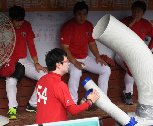 야구 유니폼을 입은 선수들이 더그아웃에 앉아 있으며, 한 선수가 대형 송풍기 앞에서 시원함을 즐기고 있다.